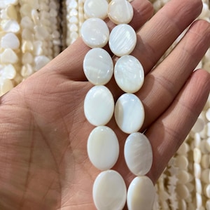 May include: A hand holds a strand of white, oval-shaped mother-of-pearl beads. The beads are smooth and have a pearlescent sheen. Other strands of similar beads are visible in the background.