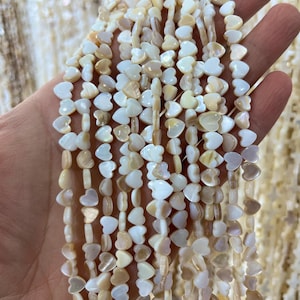 May include: Strands of heart-shaped beads in shades of white and cream are held in a hand. The beads have a pearlescent sheen and are strung together. The background shows more strands of similar beads.
