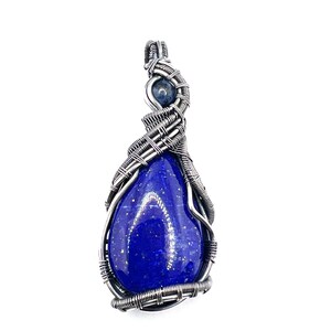 May include: A handcrafted pendant featuring a teardrop-shaped, deep blue lapis lazuli stone with gold flecks. The stone is encased in a silver-coloured wire wrap, with a smaller blue stone at the top. The pendant hangs from a silver-coloured bail.