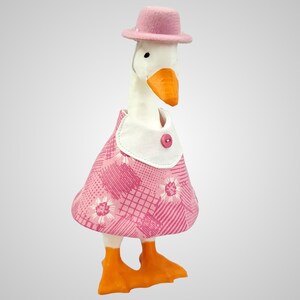 May include: A whimsical goose figurine wearing a pink dress with a white collar and a matching pink hat. The dress features a floral pattern, and the goose has orange feet and beak. The figurine is designed to stand upright.