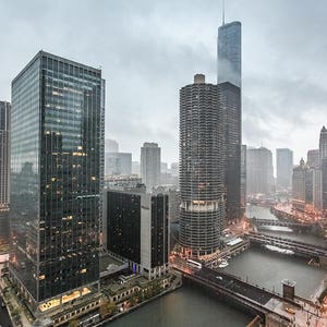 May include: A view of a city skyline with tall buildings, a river, and a bridge. The buildings are mostly gray and brown, and the river is a dark blue. The sky is overcast and gray.