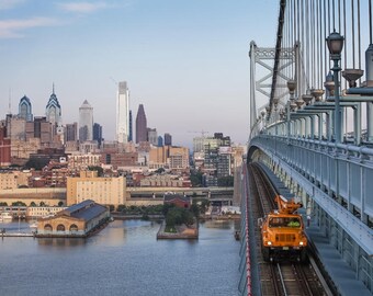 May include: A view of the Philadelphia skyline from the Benjamin Franklin Bridge. A bright orange utility truck is driving on the bridge's roadway.