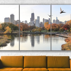 May include: A panoramic view of a city skyline with tall buildings reflected in a lake. The lake is surrounded by trees with fall foliage in shades of orange, yellow, and red. Two birds fly overhead.