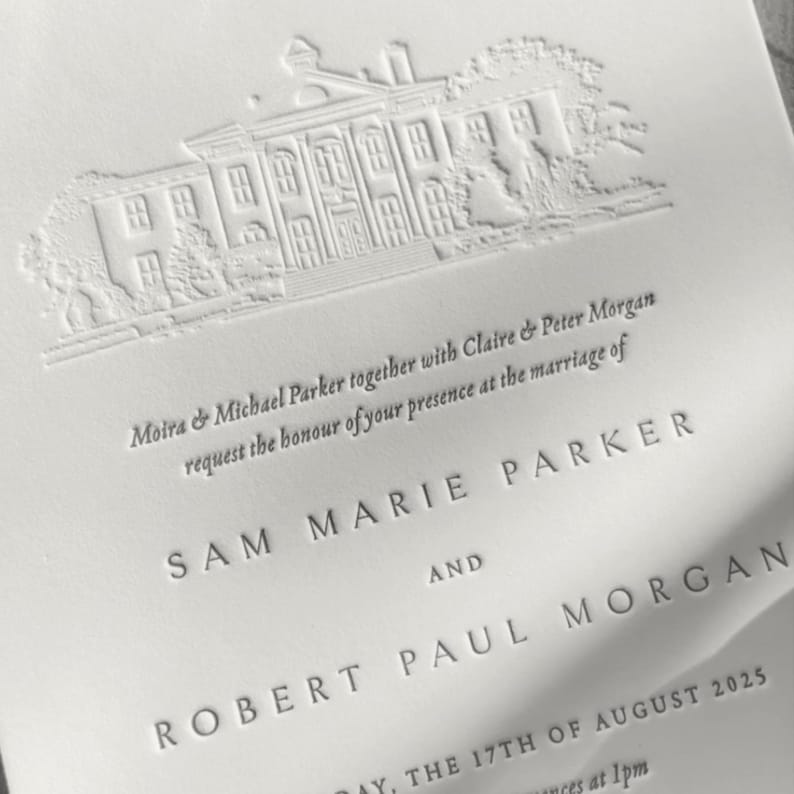 A close-up of the letterpress impression and texture on a wedding invitation with black ink letterpress text in a modern minimal and timeless serif font with a custom illustration of a wedding venue blind embossed/debossed showing texture on the thick white cotton card.