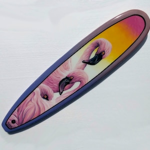 May include: A purple and pink surfboard with a painted design of three pink flamingos with black eyes and beaks.