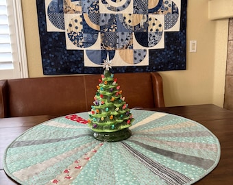 May include: A ceramic Christmas tree with colorful lights sits on a round, quilted table topper. The topper features a radial design with various fabrics in shades of blue, gray, and red. A large, blue and white quilt hangs in the background.