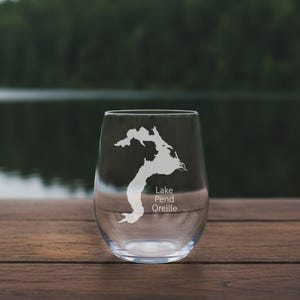 May include: A clear stemless wine glass with a white etched map of Lake Pend Oreille. The lake's name is etched below the map. The glass sits on a wooden surface with a blurred lake and trees in the background.