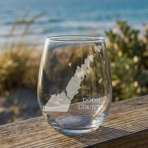 May include: A clear stemless wine glass featuring a white etched map of the Door County peninsula. The glass has a smooth, rounded silhouette and includes the text DOOR COUNTY printed in a clean, sans-serif font below the geographic outline.
