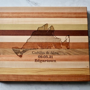May include: A wooden chopping board with a map of Long Island, New York, carved into the surface. The board is made of multiple wood types, creating a striped pattern. The text "Caitlin & Alex 09.05.21 Edgartown" is engraved below the map.