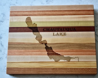 Puede incluir: Una tabla de cortar de madera con un mapa del lago Chautauqua tallado en ella. La tabla está hecha de varios tipos de madera, creando un patrón a rayas. El texto "Chautauqua Lake" está grabado en la tabla.