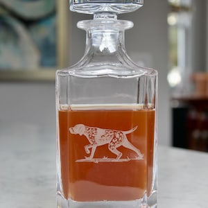 May include: A clear glass decanter with a square body and a stopper. The decanter is filled with a dark amber liquid. A white silhouette of a dog is etched into the side of the decanter.