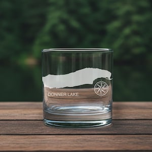 May include: A clear glass tumbler featuring an etched white silhouette of Donner Lake. The design includes the text DONNER LAKE printed in a clean sans-serif font and a detailed nautical compass rose icon positioned to the right of the lake map.