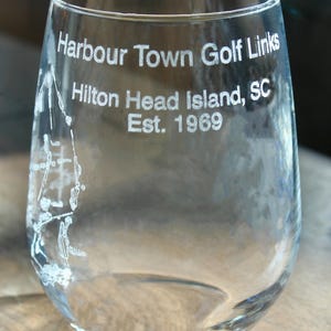 Könnte beinhalten: Klares, stieloses Weinglas mit geätztem Text: "Harbour Town Golf Links, Hilton Head Island, SC, Est. 1969." Auf der Seite ist eine Golfplatzkarte geätzt. Das Glas steht auf einer Holzoberfläche.