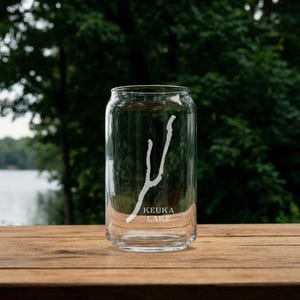 May include: A clear glass can-shaped drinking glass with the words "Keuka Lake" and a white graphic of the lake's shape. The glass sits on a wooden surface with a blurred green background.