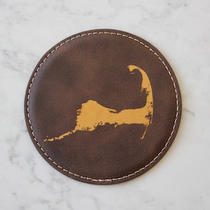 Op de afbeelding: Een bruine leren onderzetter met een gouden omtrek van Cape Cod.