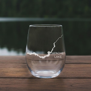May include: A clear stemless wine glass featuring a white etched map of Apache Lake. The glass has a rounded, tapered body and a smooth rim. The text "APACHE LAKE" is printed in white capital letters below the detailed map graphic on the glass.