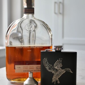 May include: A clear glass bottle of Woodford Reserve Kentucky Straight Bourbon Whiskey with a black flask featuring a gold engraved image of a duck. The bottle label reads "Woodford Reserve Kentucky Straight Bourbon Whiskey" and "45.2% ALC/VOL (90.4 PROOF)".