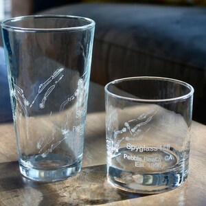 May include: Two clear drinking glasses with etched designs. One is a tall pint glass, and the other is a shorter rocks glass. Both feature a golf course layout with the text "Spyglass Hill Pebble Beach CA Est. 1966."