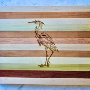 Puede incluir: Una tabla de cortar de madera con un diseño a rayas en varios tonos de marrón, beige y verde. Una ilustración detallada de una garza está grabada en el centro de la tabla.