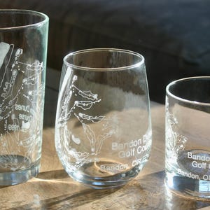 May include: Three clear glass tumblers with etched designs and text. The glasses vary in shape: a tall pint glass, a rounded stemless wine glass, and a short rocks glass. Each glass features a golf course map and the text "Bandon Dunes Golf Course, Bandon, OR."