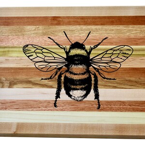 Può includere: Un tagliere in legno con un design a strisce in tonalità di marrone, beige e giallo. Un'illustrazione in bianco e nero di un'ape è centrata sul tagliere.
