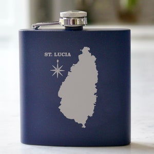Könnte beinhalten: Eine marineblaue Edelstahl-Taschenflasche mit silbernem Verschluss und einer silbernen, gravierten Karte von St. Lucia und einem Kompass.