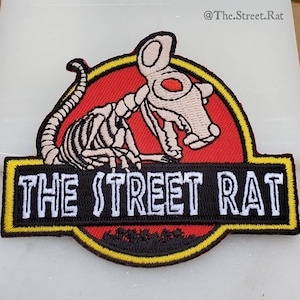 May include: A red, yellow, and black embroidered patch featuring a skeleton rat in a dinosaur pose. The text "THE STREET RAT" is written in white on a black background.