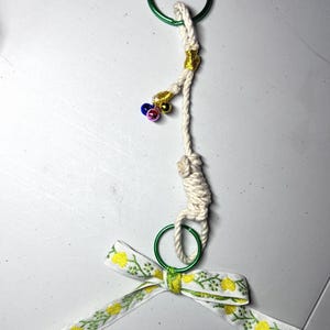 May include: A handmade keychain featuring a green metal ring, a knotted rope design, and a decorative bow. The bow is white with a yellow floral pattern. Small gold, blue, and pink bells add a touch of whimsy.