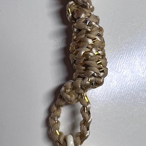 May include: A keychain with a heart-shaped pendant. The pendant is off-white with a dark brown stripe. The keychain has a gold-colored ring and a braided cord with gold accents.