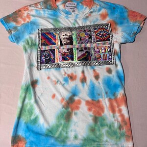 May include: A short-sleeve t-shirt with a tie-dye pattern in blue, orange, and green. The chest features a rectangular grid of six distinct graphic patches framed by silver metallic trim. The inner neck label reads INSIDE THE MAGPIE'S NEST.