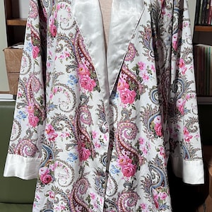 May include: A white paisley robe with a floral pattern featuring pink roses and blue accents. The robe has a satin collar and cuffs, and a button-down front. The long-sleeved robe is displayed on a mannequin.