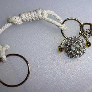 May include: A gold-toned keychain with a white rope loop and a gold ring. The keychain features a cluster of clear rhinestones, small gold charms, and a decorative key. The design includes a small key and a tiger's eye charm.