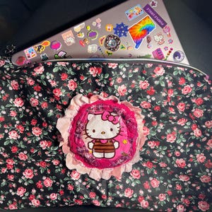 May include: A black floral print zippered pouch with a pink and white Hello Kitty applique. The pouch is filled with a laptop covered in stickers. The floral pattern features pink and red roses on a black background.