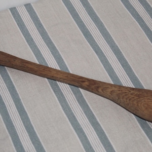 May include: A wooden muddler with a long handle and a flat, rounded end. The muddler is made of a light brown wood and rests on a striped fabric background. This bar tool is used for mixing cocktails.