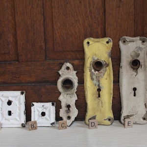 May include: An assortment of antique door hardware, including white and yellow backplates, and ornate doorknob plates. The pieces show signs of wear, with peeling paint and aged metal. The items are arranged against a wooden background.