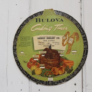 May include: A vintage Bulova Cooking Timer with a green background and black lettering. The timer features a circular dial with cooking times for various foods, including roast, steak, and vegetables. It also includes a picture of a roast with vegetables and two Bulova watches.