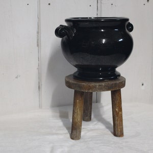 May include: A black ceramic planter with ornate handles sits on a wooden stool with three legs. The stool is made of natural wood and has a rustic finish.