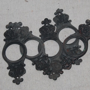 May include: A dark, weathered metal object featuring four circular openings arranged in a cluster. The piece is decorated with ornate, raised relief patterns including crown-like motifs and floral elements, with a matte, aged finish throughout.