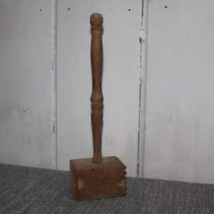 May include: A vintage wooden mallet with a square head and a long, turned handle. The mallet is a warm brown color and shows signs of wear, suggesting it's an antique tool. The handle has decorative detailing.