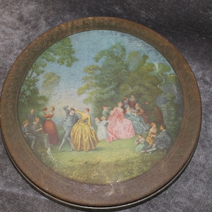 May include: A round, metal tin with a brown rim and a painted scene of people in a garden. The scene features a group of people in 18th-century clothing, including a couple dancing in the center.