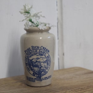 May include: A vintage cream-colored ceramic jar with blue text and a graphic. The jar is filled with white flowers. The text on the jar reads "PURE RICH CREAM STRATHBOGIE DAIRY HUNTLY".