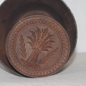 May include: A round, wooden butter mold with a wheat sheaf design. The mold is brown and sits inside a larger, dark brown container. The wheat design is carved into the wood.