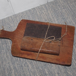 May include: A rustic wooden cutting board with a handle, featuring a rectangular block of wood and a darker, rectangular metal piece. The items are tied together with twine. The board is brown.