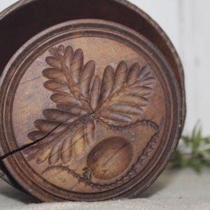 May include: A round, wooden butter mold with a detailed carved design of a leaf and a nut. The mold is brown and has a circular shape with a lid. The design is centered on the lid, and the wood has a natural grain.