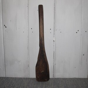 May include: A long, dark brown wooden spoon with a wide, flat paddle-shaped end and a narrow handle. The spoon is standing upright against a white wooden background.