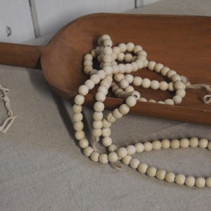 May include: A wooden scoop with a long handle holds a strand of cream-colored wooden beads. The beads are strung on a natural-colored cord. The scoop is a warm brown color.