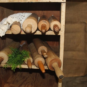 May include: A wooden shelf with multiple wooden rolling pins stored inside. The shelf is made of light wood and has a white doily on the top shelf.