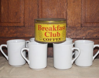 Op de afbeelding: Een gele koffieblik met de tekst "Breakfast Club Coffee" erop, omringd door zes witte keramische mokken.