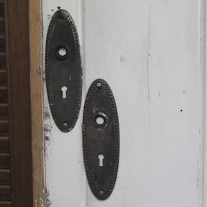 May include: Two antique oval-shaped bronze door plates with keyholes and decorative edges. The plates are mounted on a white wooden surface, with a wooden shutter visible on the left side of the image.