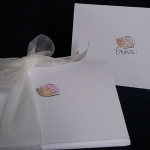 May include: A stack of white note cards with a seashell illustration and the name "Emma" on the top card. The cards are tied together with a white ribbon.